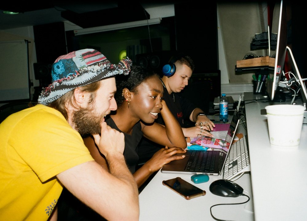 Three students are sat at music production workstations with headphones on, two are working together and smiling