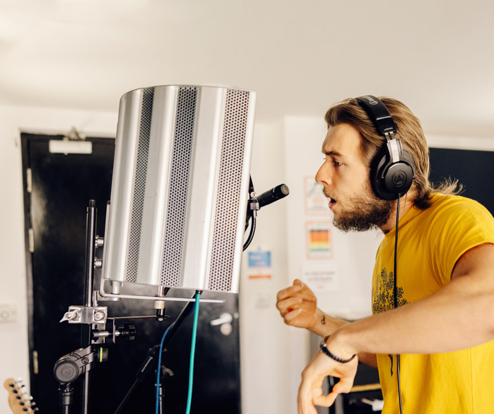 A young man in his 20s is in the vocal booth of a music studio, he is midway through rapping into the microphone
