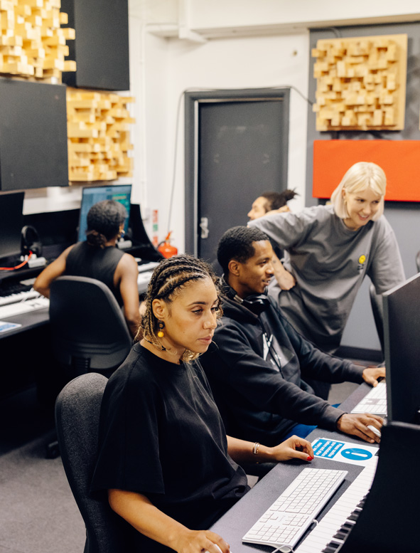 Four students are sat in the music production training room with a tutor overseeing their work and smiling